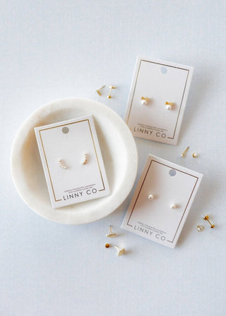 Gold earrings on LINNY CO cards with a marble dish on a light gray background