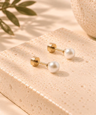 A pair of gold post earrings with round white pearl studs, displayed on a textured beige stone block with soft shadows and a leaf in the background.
