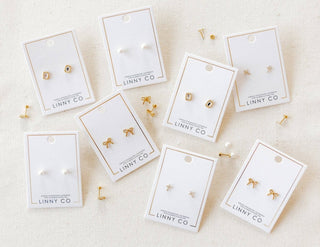 Gold earrings on LINNY CO branded cards on a beige background