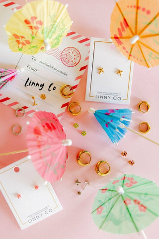 A styled flat-lay of small gold jewelry pieces, including star and heart stud earrings and ring charms, displayed on white cards with cocktail umbrellas and a postcard design on a pink background. The brand name LINNY CO is visible on the cards.
