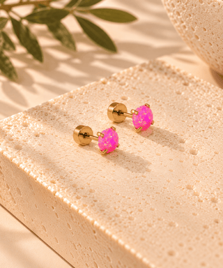 A pair of gold stud earrings with vibrant pink iridescent cabochon stones, displayed on a textured beige stone block with soft shadows and green leaves in the background.