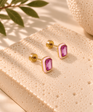 A pair of gold-toned stud earrings with rectangular pink bezel-set purple gemstones, displayed on a textured beige stone slab with soft shadows and a leaf in the background.