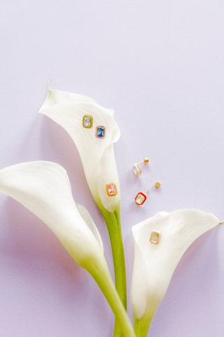 Gold earrings with gemstones on a white flower against a light purple background
