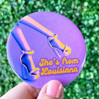 Button badge with high heels and 'She's from Louisiana' text held against a green leafy background