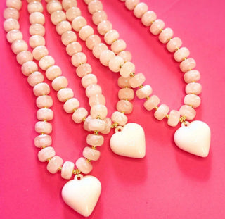 Three beaded necklaces with pale pink round beads and white heart-shaped pendants, arranged on a bright pink background.
