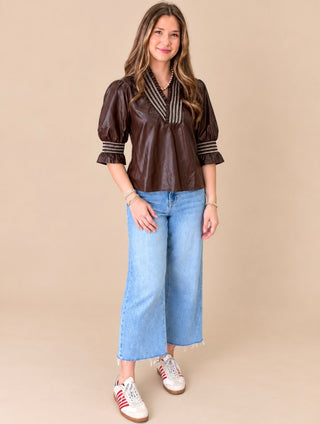 A woman modeling a dark brown faux-leather puff-sleeve blouse with striped trim on the V-neck and cuffs, paired with light blue wide-leg jeans and white sneakers, against a solid beige studio background.