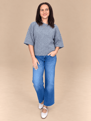 A woman modeling a heathered blue short-sleeve knit top and blue jeans against a solid beige studio background.