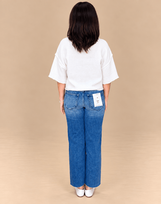 A woman viewed from behind wearing a white short-sleeve top and blue wide-leg jeans, standing against a plain beige studio background.