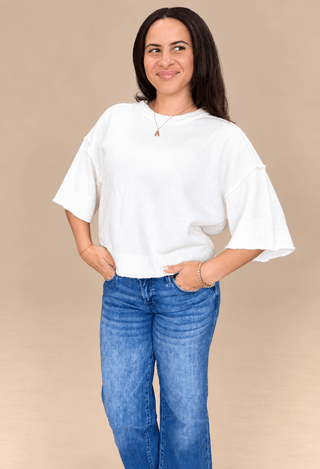 A woman modeling a white short-sleeve knit top with flared bell sleeves and blue jeans, standing against a solid beige studio background.