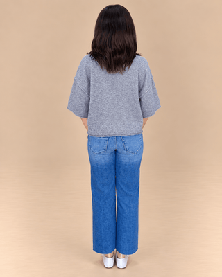 A woman viewed from behind wearing a gray short-sleeve knit top and blue wide-leg jeans, standing against a plain beige studio background.