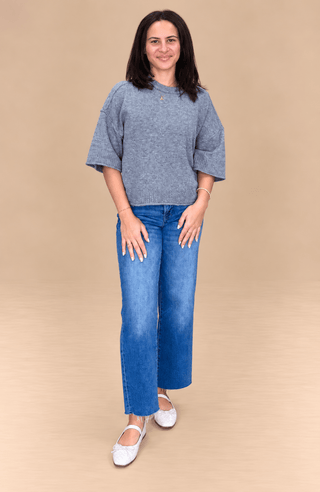 A woman modeling a heather gray short-sleeve knit top and blue jeans against a solid beige studio background.