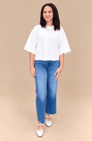 A woman modeling a white short-sleeve top and blue jeans against a solid beige studio background.