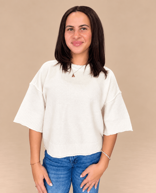 A woman with dark shoulder-length hair wearing a cream-colored knit sweater with wide, flared three-quarter sleeves and blue jeans, standing against a solid beige background.