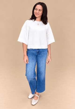 A woman modeling a white short-sleeve knit top and blue wide-leg jeans against a solid beige studio background.