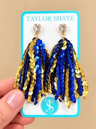 A pair of gold and blue sequin tassel earrings on a white display card with the brand name Taylor Shaye, held by a hand against a beige background.