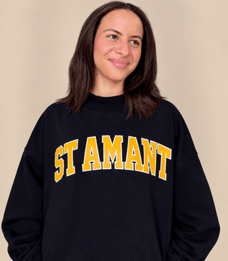 A woman wearing a black crewneck sweatshirt with 'ST AMANT' printed in yellow varsity-style letters with a white outline across the chest, set against a plain beige background.