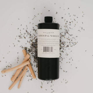 A bottle of HOLLYWOOD Ashwood & Mahogany room spray with a black body and white label, styled on a white background with scattered ashwood chips and wooden sticks.