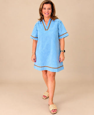 A woman modeling a light blue short-sleeve tunic-style dress with brown trim details, standing against a solid beige studio background.