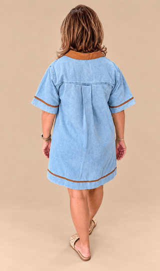 A woman viewed from behind wearing a light blue denim tunic dress with brown trim on the collar, sleeve cuffs, and hem, paired with tan slide sandals, against a solid beige studio background.