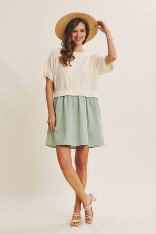 A woman modeling a two-piece outfit consisting of a cream short-sleeve knit top and a sage green pleated mini skirt, styled with a straw hat and white heeled sandals against a plain cream studio background.