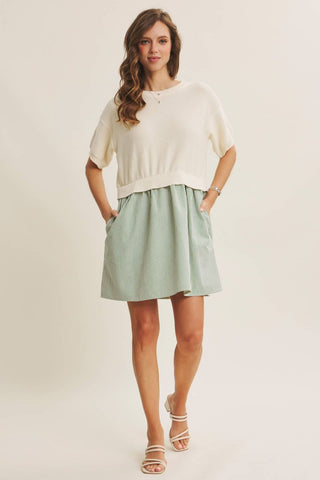 A woman modeling a cream short-sleeve knit top and a light sage green mini skirt with side pockets, paired with white strappy sandals, against a plain cream studio background.
