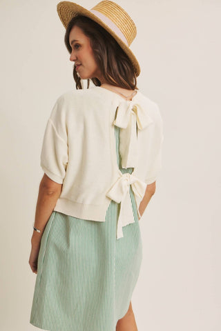A woman wearing a cream knit short-sleeve top with a large bow detail at the back and a light green and white striped skirt, shown from the back against a plain light beige background.