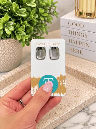 A hand holds a white jewelry display card featuring a pair of square stud earrings with iridescent purple-blue stones and silver settings. The card is branded Taylor Shaye, with a gold logo and gold brushstroke design near the bottom.