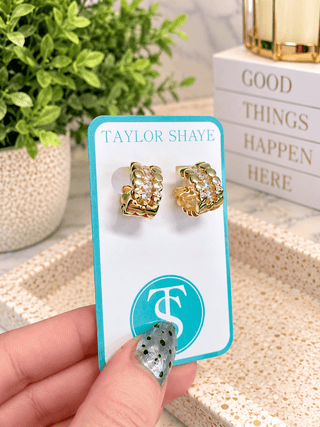 A pair of gold huggie hoop earrings with clear crystals, displayed on a turquoise and white Taylor Shaye card held by a hand. The background features a potted plant and a stack of books.