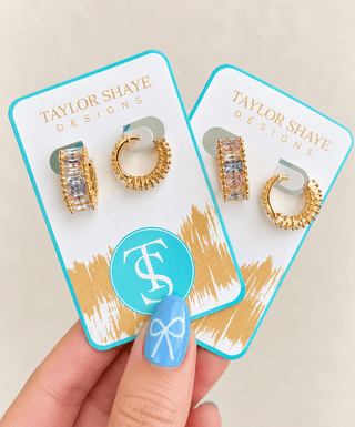 A hand with blue nail art holds two gold hoop earrings with rectangular pavé crystals on branded Taylor Shaye Designs display cards against a light beige background.