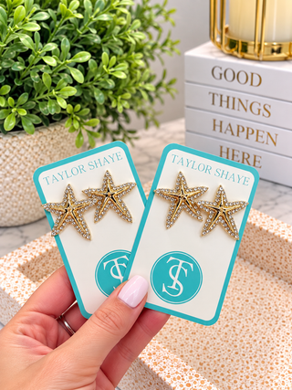 A hand holds two turquoise and white jewelry display cards, each featuring a pair of gold starfish stud earrings with rhinestone-like detailing. The cards are branded Taylor Shaye, and the background shows a potted plant, books, and a decorative tray.