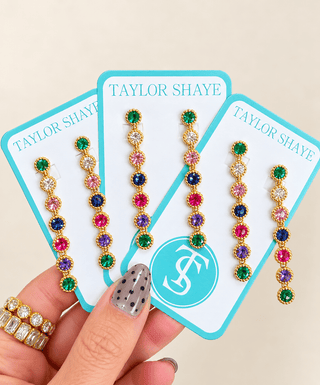 Three pairs of dangling gold-tone earrings with colorful gemstones are displayed on branded Taylor Shaye cards, held by a hand with nail art and rings against a light beige background.