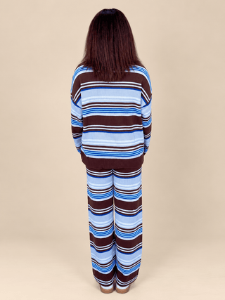 A woman viewed from behind wearing a matching two-piece knit loungewear set with horizontal stripes in brown, light blue, navy, and white, set against a solid beige background.