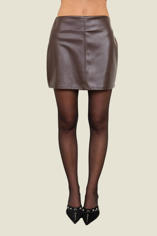 A model's lower body wearing a brown faux-leather mini skirt, sheer black tights, and black studded heels, standing against a plain off-white wall.