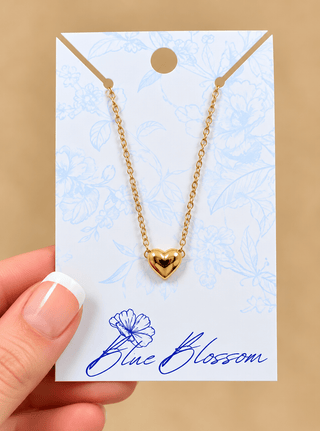 A gold heart pendant necklace on a delicate gold chain, displayed on a white card with blue floral patterns and the brand name 'Blue Blossom'. A hand with a French manicure is holding the card on the left side, set against a solid beige background.