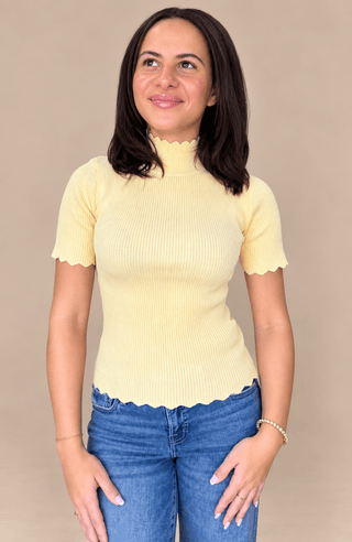 A woman wearing a pale yellow short-sleeve ribbed mock neck top with scalloped edges and blue jeans, standing against a solid beige background.