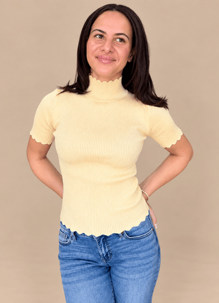 Model wearing a fitted pastel yellow mock neck knit tee with scalloped edges and jeans with a tan background.