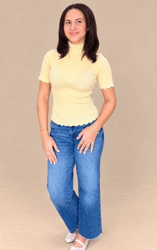 A woman modeling a pale yellow short-sleeve mock neck top with scalloped edges and blue wide-leg jeans, standing against a solid beige studio background.