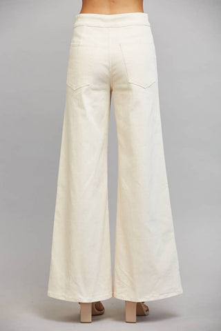 Back view of a person wearing off-white wide-leg pants with a high waistband and back patch pockets, paired with beige block-heel sandals against a light gray studio background.