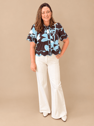 A woman modeling a short-sleeve button-up blouse with a brown, light blue, and white floral pattern, paired with white wide-leg pants, against a solid beige studio background.