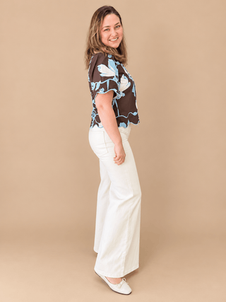 A woman modeling a sheer brown short-sleeve blouse with a blue and white floral pattern, paired with white wide-leg pants and white flats, against a solid beige studio background.