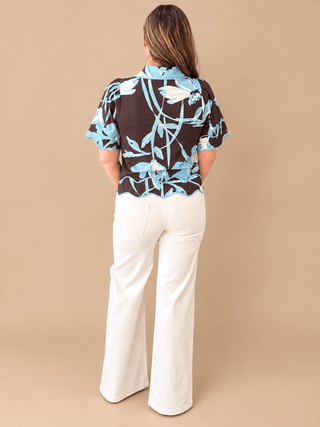 A woman viewed from behind wearing a short-sleeve button-up blouse with a brown, light blue, and white floral pattern, paired with white wide-leg pants, against a solid beige studio background.