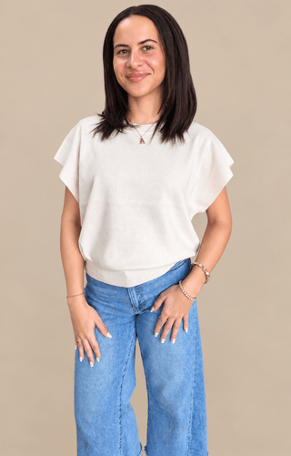 A woman modeling a cream-colored short-sleeve knit top with ruffled edges, paired with blue jeans, against a solid beige background.