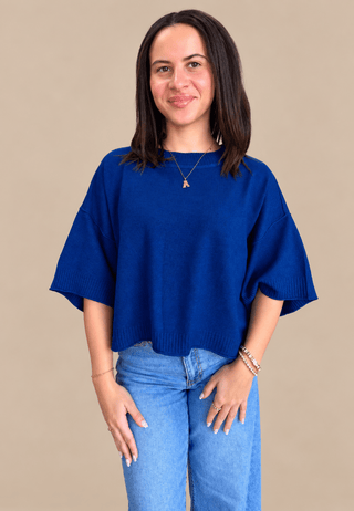 A woman wearing a vibrant royal blue knit top with wide, short sleeves and light wash blue jeans, standing against a solid beige background.