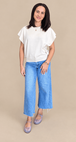 A woman modeling a white short-sleeve top and wide-leg blue jeans against a solid beige studio background.
