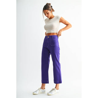 A woman modeling a pair of bright purple cropped wide-leg jeans with a white ribbed crop top and white sneakers, standing against a plain white studio background.