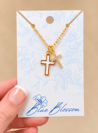A gold cross necklace with a small crystal-embellished cross pendant, displayed on a decorative white card held by a hand. The card features a light blue floral pattern and the brand name 'Blue Blossom' printed at the bottom.