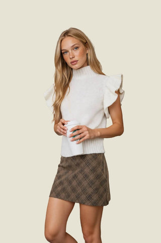 A woman stands indoors against a stone wall wearing a white mock-neck top with ruffled cap sleeves and a brown plaid mini skirt, holding a white coffee cup.