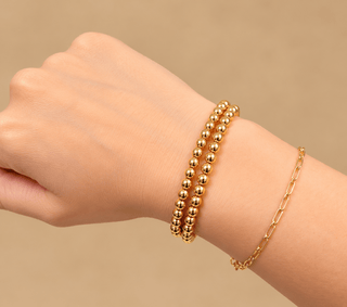 A close-up of a person's wrist wearing two gold bracelets: one with small metallic beads and one with a delicate chain link design, against a soft beige background.