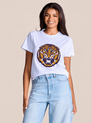 A woman wearing a white short-sleeve t-shirt with a large embroidered tiger head graphic in purple, yellow, and orange, paired with light-wash blue jeans, against a solid beige background.