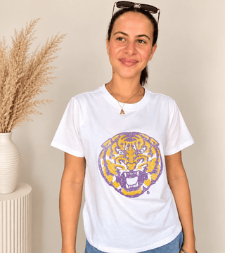 A woman wearing a white short-sleeve t-shirt with a distressed purple and yellow tiger graphic on the front, standing indoors next to a vase of dried grass.